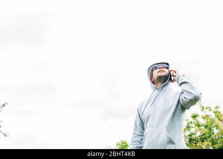 Junger Mann in grauem Sweatshirt und blauer Sonnenbrille, der das Handy in seinen Händen hält und nach oben schaut Stockfoto