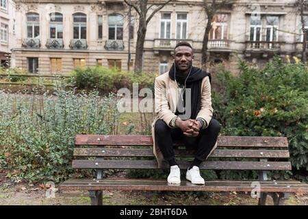 Junge schwarze bedeuten, tragen beigefarbenen Mantel auf der Rückenlehne der Parkbank mit Blick auf die Kamera sitzen. Stockfoto