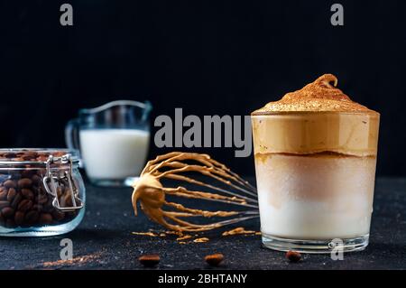 Iced Dalgona Kaffee mit Corolla, Milch und Kaffeebohnen auf dunklem Hintergrund. Trendy Cremigen Schlagsahne Kaffee. Südkoreanischer Kaltsommer-Drink. Stockfoto