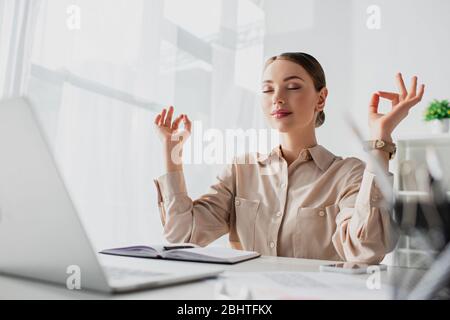 Schöne Geschäftsfrau meditiert mit geschlossenen Augen und Gyan Mudra am Arbeitsplatz mit Laptop Stockfoto