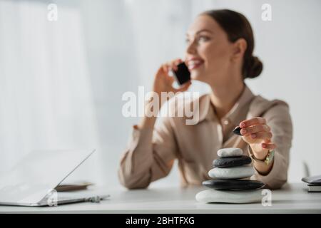 Selektiver Fokus der Geschäftsfrau, die am Arbeitsplatz mit Zen-Steinen auf dem Smartphone spricht Stockfoto