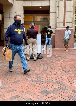 Reston, VA, USA -- 25. April 2020. Ein Mann, der eine Gesichtsmaske trägt, trägt sein Essen zum Mitnehmen in einer Tasche; andere warten, bis ihre Befehle erfüllt sind. Stockfoto