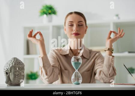 Selektiver Fokus der Geschäftsfrau, die mit geschlossenen Augen und Gyan Mudra am Arbeitsplatz meditiert, mit Buddha-Kopf, Sanduhr und Räucherstäbchen Stockfoto