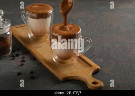 Dalgona Kaffee, ein trendiges Getränk. Zwei Gläser Dalgona Kaffee auf einem Holzbrett, neben Kaffeebohnen und Instantkaffee in einem Glasgefäß. Vertikale ausrichtung Stockfoto