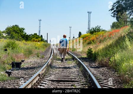 Ein älterer Glatzmann geht auf den Bahngleisen in die Ferne, vorbei an einem Damm mit Mohnblumen gegen einen blauen Himmel. Flucht aus der Stadt und sich selbst. Stockfoto