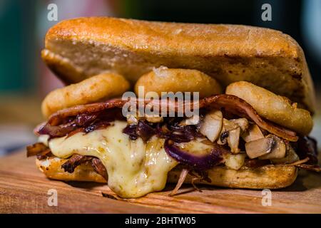 Rustikales Sandwich aus zerfetztem Fleisch mit Käse, Zwiebelringen, gebratenem Speck und Pilzen, auf einem Holzbrett Stockfoto