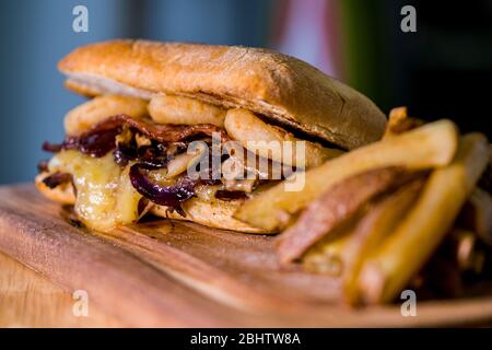 Rustikales Sandwich aus geschreddertem Fleisch mit Käse, Zwiebelringen, gebratenem Speck und Pilzen, begleitet von pommes frites, auf einem Holzbrett Stockfoto