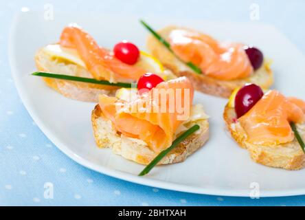 Brot in Scheiben mit Butter und Lachs, dekoriert mit Zitronenscheibe, Preiselbeere und grüner Zwiebel auf weißer Schale Stockfoto