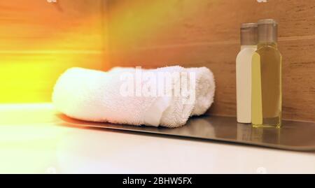 Rollen Sie ein weißes Handtuch auf einem Tisch in einem Spa-Hotel-Badezimmer auf unscharfer Holzuntergrund mit Sonnenstrahlen Stockfoto