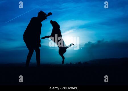 Silhouette eines jungen Mannes mit seinem Hund springt mit blauem Nachtlicht Stockfoto