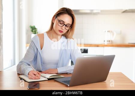Frau arbeitet auf Laptop zu Hause in der Küche. Schreiben auf Notizblock. Geschäftsfrau, die über etwas wichtiges nachdenkt. Frau, die versucht, einen Job zu finden. Crysis Stockfoto