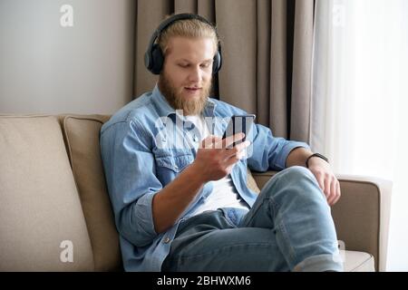 Junger Mann, der Kopfhörer trägt und das Telefon benutzt, um zu Hause auf den Bildschirm zu schauen. Stockfoto