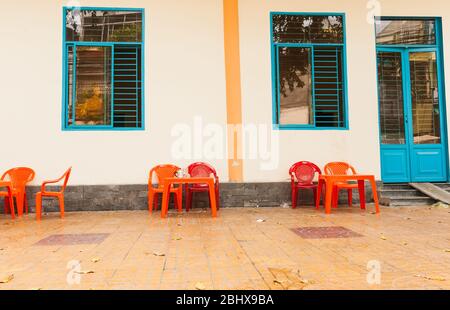 Leere Sätze von roten und orangefarbenen Stühlen und Tischen auf dem Fußweg aus dem Seitengebäude mit blauen Türen und Fenstern in der Stadt Can Tho, Vietnam. Stockfoto