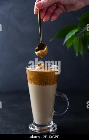 Dalgona Kaffee zubereiten. Hand mit einem Löffel Schlagschaum auf schwarzem Hintergrund Stockfoto