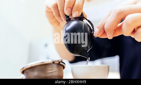 Nahaufnahme der Hände, die chinesischen grünen Tee aus dem kleinen Keramikkessel in eine kleine Tasse mit Teesieb im Vordergrund gießen. Stockfoto