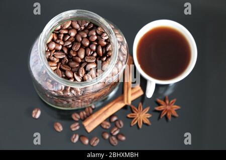 Kaffee mit Gewürzen, dunkel gerösteten Kaffeebohnen im Glasgefäß, Sternanis, Zimtstangen und weiße Tasse auf dunklem Glastisch Stockfoto