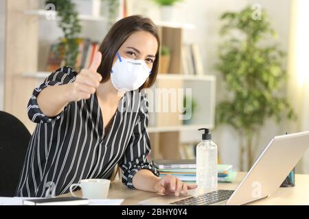 Frau mit Daumen hoch, die Schutzmaske trägt Telearbeit mit Laptop in Coronavirus-Quarantäne, die auf einem Schreibtisch im Homeoffice sitzt Stockfoto