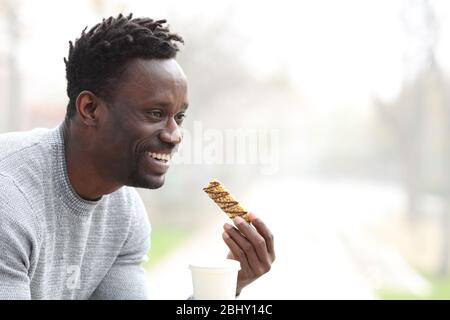 Ein glücklicher schwarzer Mann, der Kaffee zum Mitnehmen und eine Müsliriegel zum Mitnehmen in einer Parkbank serviert hat Stockfoto