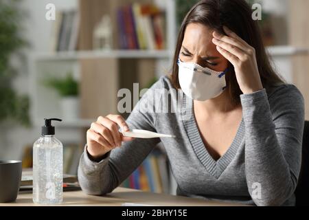 Kranke Frau mit Schutzmaske leidet Coronavirus Symptome mit Kopfschmerzen hält Thermometer in der Nacht zu Hause Stockfoto