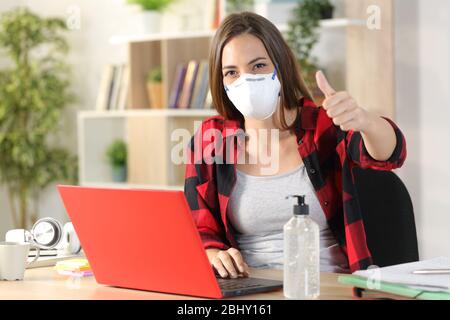 Studentin Frau mit Schutzmaske due covid-19 Gesturing Daumen hoch aussehende Kamera mit Laptop sitzt auf einem Schreibtisch zu Hause Stockfoto