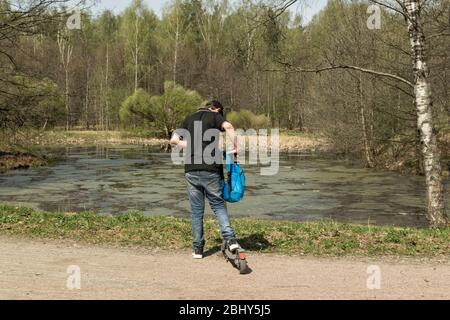 Der junge Mann in einem schwarzen T-Shirt, mit Rucksack und Elektroroller in einem Park in der Nähe eines Waldsees steht zurück Stockfoto