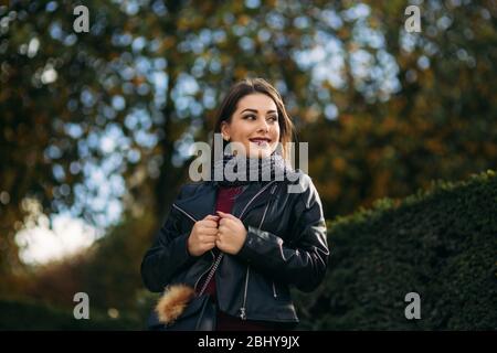 Stylische schöne Dame in einer schwarzen Lederjacke mit schwarzer Tasche und burgunderfarbener Bluse. Attraktive junge Frau lächeln Stockfoto