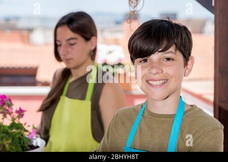 Junger Teenager trägt eine Gärtnerschürze, während er auf der Terrasse zu Hause steht und fröhlich zu einer Kamera lächelt. Stockfoto