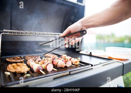 Hand des jungen Mannes Grillen einige Fleisch-und Gemüse-Fleisch-Spieße auf riesigen Gasgrill, BBQ-Konzept Stockfoto