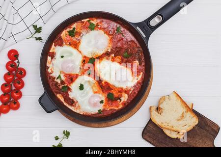 Shakshuka Eiergericht mit Tomatensauce in einer schwarzen Gusseisenpfanne auf dem weißen Holztisch Stockfoto