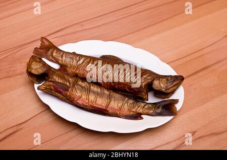 Zwei geräucherte Fische, Bachforelle, auf einem weißen Teller auf einem Buchentisch. Stockfoto