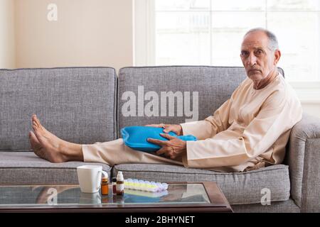 Älterer Mann sitzt auf dem Sofa massiert seine Knie mit einem heißen Wasserbeutel. (Gesundheit und Fitness) Stockfoto