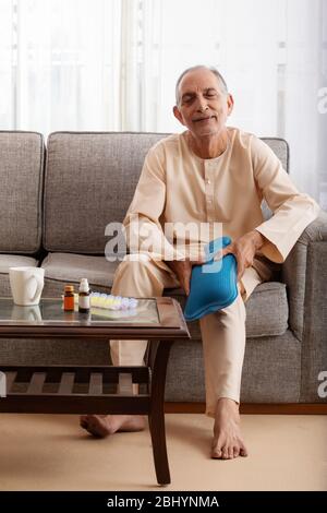 Älterer Mann sitzt auf dem Sofa massiert seine Knie mit einem heißen Wasserbeutel. (Gesundheit und Fitness) Stockfoto