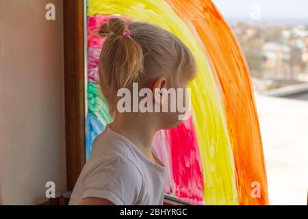 Kleines Mädchen malte einen Regenbogen auf eine Fensterscheibe Nahaufnahme. Regenbogen auf dem Glas Flash Mob. Quarantäne des Coronavirus. Familienisolierung im Innenbereich. Mädchen während Stockfoto