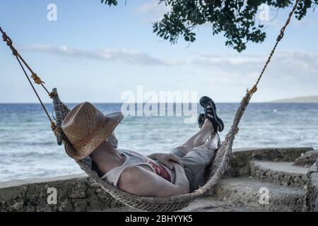 Ein Mann genießt Ruhe, liegt in einer Hängematte auf dem Hintergrund des Ozeans und Sonnenuntergang unter einem Baum. Stockfoto