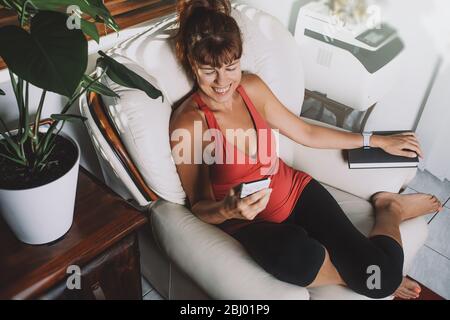 Charmante Frau mittleren Alters, die auf das Smartphone schaut und lächelt, mit einem Buch sitzt auf einem Sessel im Wohnzimmer. Stockfoto