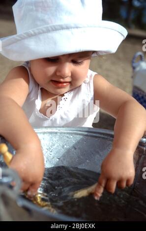 Kleines Mädchen spielt mit einem Eimer mit klarem Wasser und einigen Wäscheklammern, während Mutter die Wäsche im sonnigen Hof hängt - Stockfoto