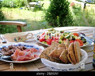 Schwedisches Sommermittagessen mit Schinken, Wurst und kaltem Fleisch und Salat mit flachem Roggenbrot, serviert auf Holztisch im Schatten der Bäume - Stockfoto