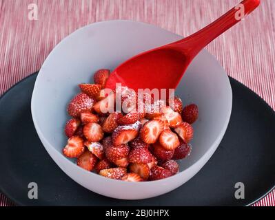 Erdbeeren, geschält und halbiert, mit Gießzucker bestäubt, in einer Schüssel aus mattem Glas auf schwarzem Teller mit hellrotem Melaminlöffel serviert. - Stockfoto