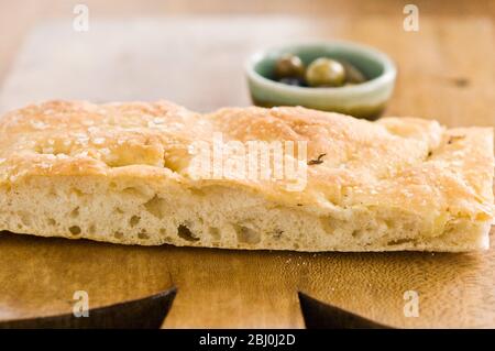 Olivenöl und Rosmarin Foccaccia Brot auf Holzbrett mit kleinen Schüsseln von einkerkerntem Olivenöl und ganze Oliven. - Stockfoto