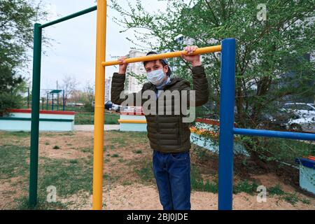 Teenager-Junge steht in der Nähe horizontale Bar von Outdoor-Trainingsgeräten, auf Spielplatz in der Nähe von Hochhäusern mit Wohnungen, eine medizinische Maske auf seinem Gesicht p Stockfoto