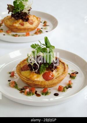 Kleine herzhafte Party Quiches mit dekorativer Beilage von Baby-Blatt-Salat und umgeben von fein gehackten Tomaten und Kräutern mit Öl und Balsamico-Rebe Stockfoto