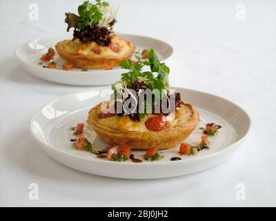 Kleine herzhafte Party Quiches mit dekorativer Beilage von Baby-Blatt-Salat und umgeben von fein gehackten Tomaten und Kräutern mit Öl und Balsamico-Rebe Stockfoto