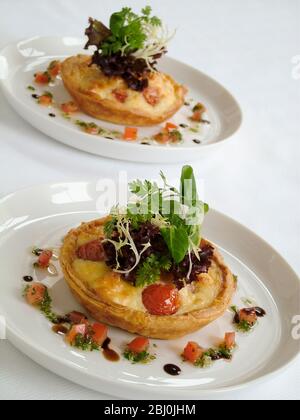 Kleine herzhafte Party Quiches mit dekorativer Beilage von Baby-Blatt-Salat und umgeben von fein gehackten Tomaten und Kräutern mit Öl und Balsamico-Rebe Stockfoto