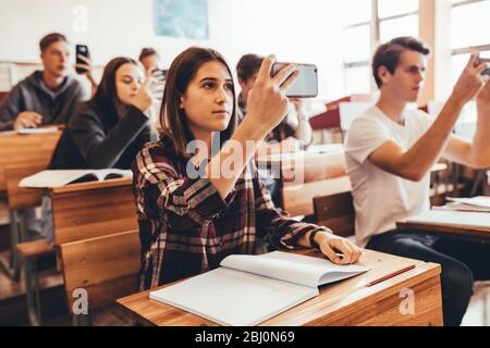 Schüler, die den Vortrag im Klassenzimmer aufzeichnen. Gruppe von Jungen und Mädchen, die den Vortrag eines Lehrers im Klassenzimmer mit ihren Mobiltelefonen Filmen. Stockfoto