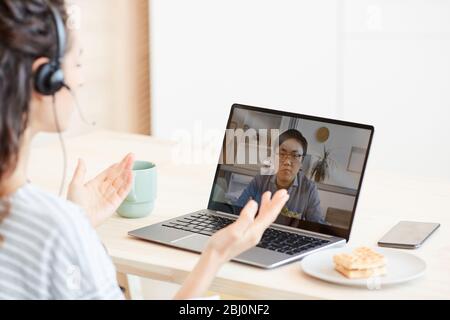 Junge Frau sitzt am Tisch in der Küche und diskutiert mit ihrem Kollegen über Videoanruf Stockfoto