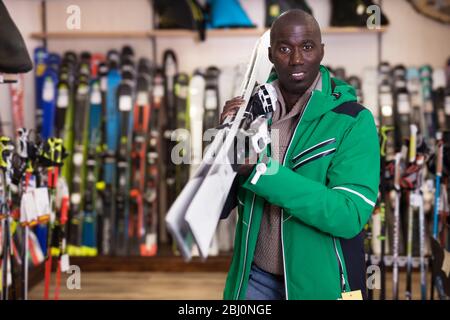 Glücklicher junger Afrikaner im Skioutfit, der mit gekaufter Skiausrüstung im Shop steht Stockfoto