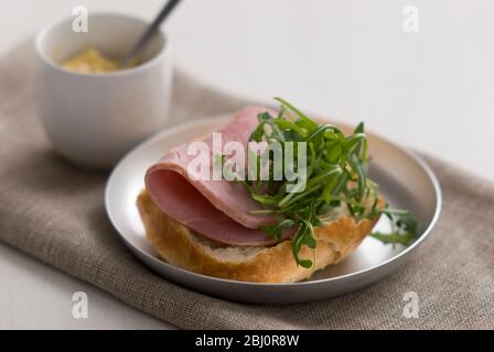 Offenes Sandwich aus knuspriger Pain Rustique Rolle, mit dicker Scheibe Qualitätsschinken und Rucola-Salatblatt-Garnitur, mit kleinem Topf Dijon-Senf - Stockfoto