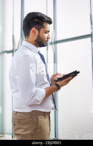 Junger Mann, der im Büro am Fenster steht und auf seinem Tablet arbeitet. Stockfoto