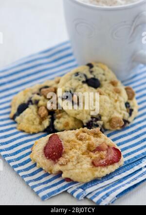 Weiche Plätzchen mit Beeren und Nüssen auf blau-weiß gestreifter Serviette mit Becher Cappucino - Stockfoto