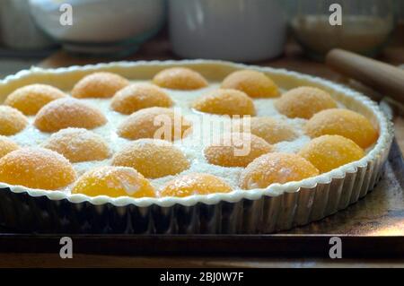 Herstellung Pfirsichtart mit halbierten Pfirsichen und Creme Patissiere auf Blätterteig in Metalldose, bestreut mit Gießzucker und gemahlenen Mandeln. - Stockfoto